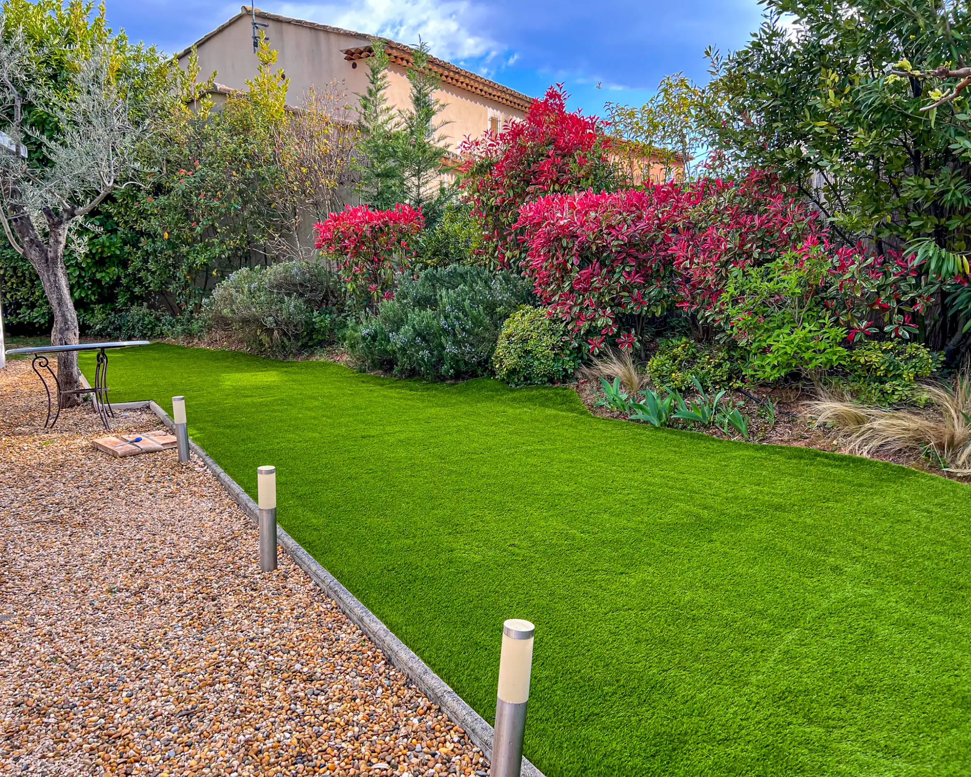 Pelouse en gazon synthétique posée à Meyreuil, délimitée par une allée de graviers et un massif de plantes colorées (Photinia) pour un jardin sans entretien.
