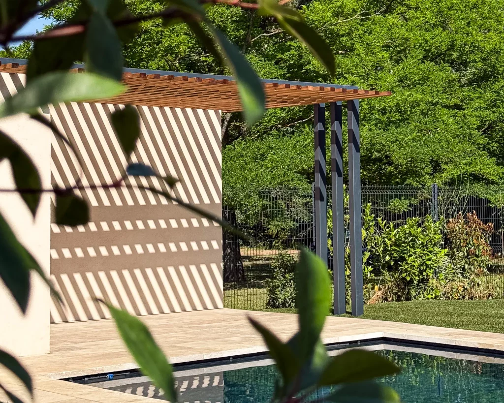 Vue artistique d'une pergola design en bois et métal, créant des jeux d'ombres graphiques sur une terrasse de piscine à Aix-en-Provence, avec la végétation en premier plan.